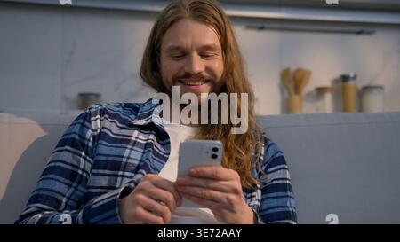 Happy smiling Caucasian man sitting on couch at home using mobile phone indoor male relaxed carefree guy scrolling social media apps smartphone Stock Photo