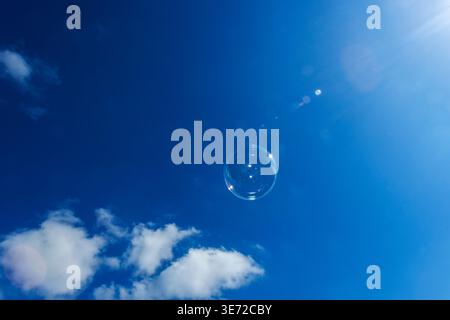 A single soap bubble against a blue sky with light clouds, backlit by the sun. Stock Photo