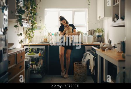 A mother cradles her baby in a sunlit kitchen filled with plants and everyday kitchenware. A tender moment of care and home life. Stock Photo