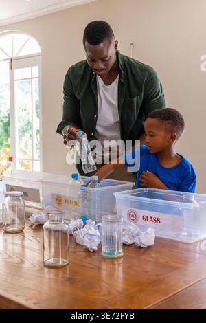 African American father and son sorting recyclables at table by arched glass door with labeled bins. Parent, child, recycling, home, daylight, wood, b Stock Photo