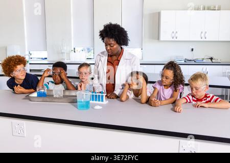African American female teacher in lab coat guiding six children testing blue liquid at school lab. Classroom, science, students, experimentation, cur Stock Photo