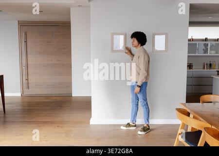 African American man standing touching two small frames on partition wall in beige sweater at home. Pictures, decor, contemporary, minimalist, interio Stock Photo