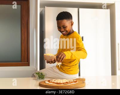 African American child wearing yellow top sprinkling cheese over pizza on countertop in kitchen. Kid, smile, prep, greens, spinach, bowl, modern Stock Photo