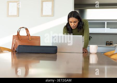 African American woman leaning over silver laptop at kitchen table with handbag, case, copy space. Workspace, daylight, home, concentration, productiv Stock Photo