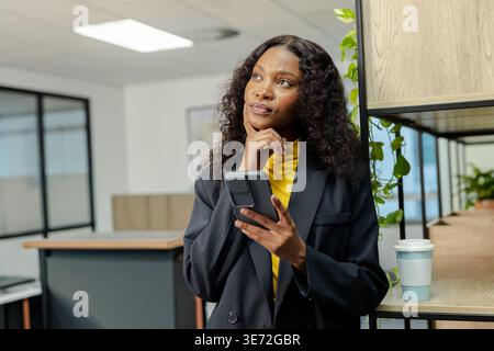 African American woman standing in office in black blazer yellow turtleneck holding phone near cup. Minimalist, bright, modern, openplan, workspace, p Stock Photo
