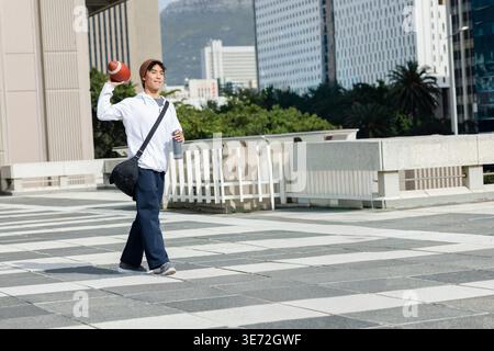 Asian man walking across tiled plaza wearing beanie holding football and duffel, copy space. Rooftop, motion, athletic, urban, crossbody, sunny, skyli Stock Photo