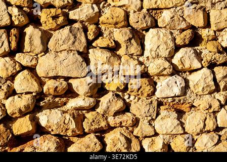 Sunlit close-up of rough limestone stones and mortar in a rustic wall at Castell Denia, showing irregular blocks and textured joints. Stock Photo