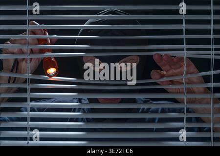 A man with a menacing expression and a balaclava controls the situation from a Venetian blind with a torch in his hand Stock Photo