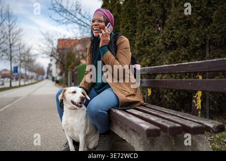 Happy woman talking on phone with dog at park bench Stock Photo