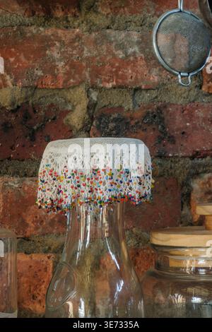 Still life with a glass bottle with an ornamental beaded doily in a rustic kitchen environment with a red brick wall. Stock Photo