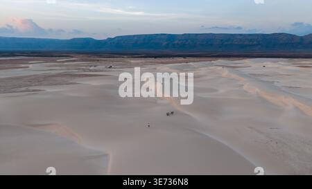 Aerial view of pale sand dunes with a few tiny figures walking along ridges, distant flat mountains under a soft pastel sky at dusk. Zahek Sand Dunes, Stock Photo