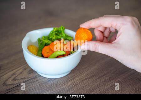 Hand taking carrot vegetable chip from bowl on wooden table. Healthy snack choice and clean eating lifestyle concept. Stock Photo