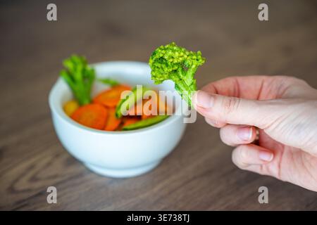 Hand holding broccoli vegetable chip above bowl on wooden table. Healthy snack alternative and clean eating lifestyle concept. Stock Photo