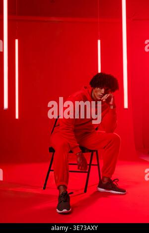 African American male leaning on black metal chair in studio wearing red tracksuit among LED tubes. Crimson, monochrome, minimalist, editorial, portra Stock Photo