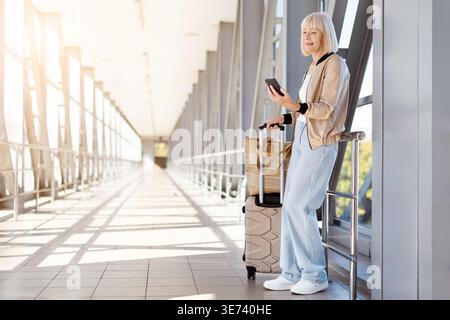 Mature woman using smartphone in airport terminal while standing with luggage Stock Photo