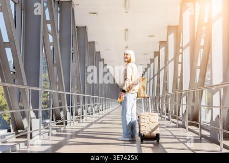 Mature woman standing in airport terminal with suitcase and passport, looking back Stock Photo