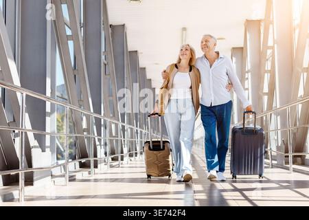 Mature couple walking together through sunlit airport terminal, pulling suitcases Stock Photo