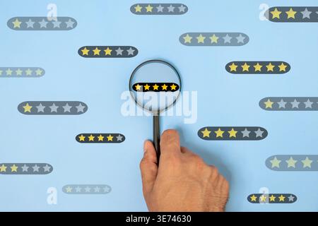 Hand holding a magnifying glass focusing on a five-star rating on a blue background. Customer feedback, quality assurance, consumer reviews concept. Stock Photo