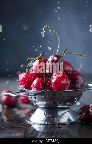 Fresh red cherries, plump and vibrant, are generously piled into a gleaming stainless steel colander. Water splashes are dynamically frozen in mid-air Stock Photo