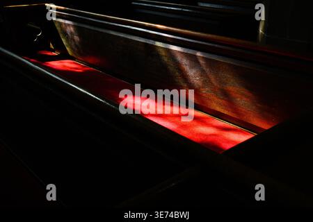 Sunlight falls on red seat and wooden back on old church pew with shadows in surrounding Stock Photo