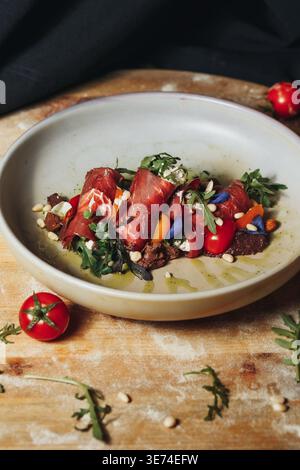 Gourmet Bresaola rolls stuffed with cream cheese and herbs served with cherry tomatoes pine nuts and fresh arugula in a ceramic bowl. Stock Photo