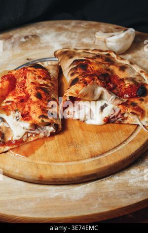Traditional Italian calzone pizza cut in half with melted mozzarella cheese ham and mushrooms on a rustic wooden board with flour. Stock Photo