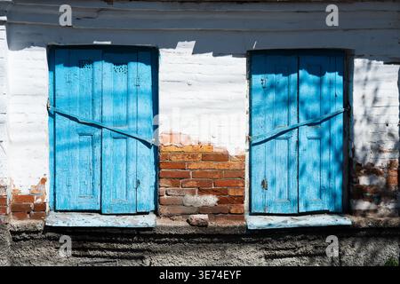 Vintage cottage exterior with peeling paint and exposed brickwork. Concept of rural life and heritage. Old white brick wall with two closed blue woode Stock Photo