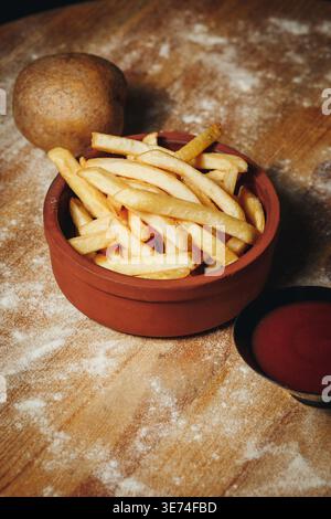 Crispy golden french fries served in a rustic clay bowl with tomato ketchup on a floured wooden table with a whole raw potato. Stock Photo