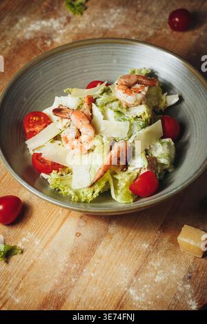Fresh Caesar salad with grilled shrimps, cherry tomatoes, parmesan shavings and crisp lettuce in a ceramic bowl on a rustic wooden table. Stock Photo