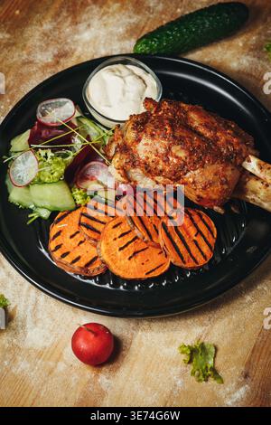 Roasted pork knuckle served with grilled sweet potato slices a fresh garden salad and creamy dipping sauce on a black plate. Stock Photo