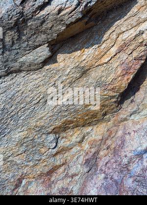 Close-up view of a rugged, steep natural rock face highlighting its complex layered textures, varied colors, and deep fissures under bright sunlight. Stock Photo