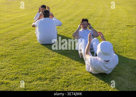 Diverse male teammates doing partner sit-ups on grassy sports field, wearing white clothes and caps. Athletes, outdoor, turf, sunlit, shadows, workout Stock Photo