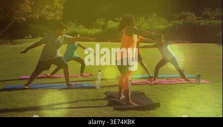 Leading instructor in orange tank guiding children doing Warrior II on mats in park, water bottles. Outdoor, yoga, leader, kids, group, sunshine, natu Stock Photo