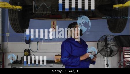 Inspecting woman tech in blue coverall mask checking tablet under vehicle on lift in workshop, data. Garage, mechanic, inspection, diagnostics, device Stock Photo