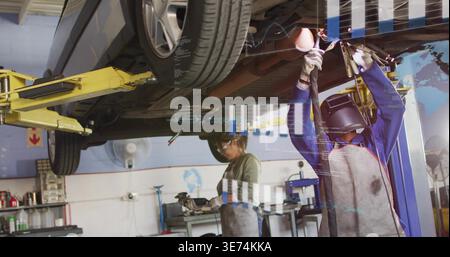 Repairing two female techs wearing helmet and aprons using grinder under lifted car in garage, AR. Technician, workshop, automotive, repair, hoist, to Stock Photo
