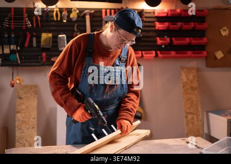 Female carpenter using cordless electric drill while working in furniture workshop. Woodworking studio. Production process. Stock Photo