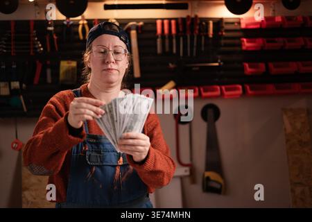 female carpenter in blue overalls counting money in her carpentry workshop. Concept of salary. copy space Stock Photo
