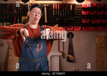 displeased carpenter woman wearing overalls in a carpentry workshop showing thumbs-down gesture. copy space Stock Photo