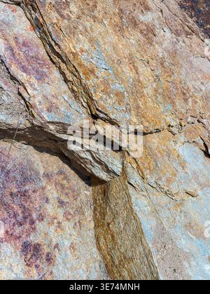 A detailed close-up of a sturdy rock face featuring a prominent diagonal crack and varying textures with patches of rust and grayish-blue minerals. Th Stock Photo