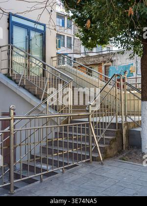 A set of outdoor concrete stairs equipped with shiny stainless steel handrails leads up to a residential building entrance on a sunny day. It depicts Stock Photo
