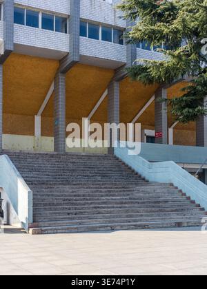 A grand, wide set of empty concrete stairs leads up to the entrance of a massive public facility, featuring tall support pillars and an orange-paneled Stock Photo