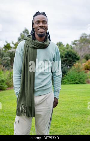 African American man 20s standing on lawn in park, wearing green sweatshirt olive scarf cargo pants. Male, outdoor, greenspace, casual, muted, neckwea Stock Photo