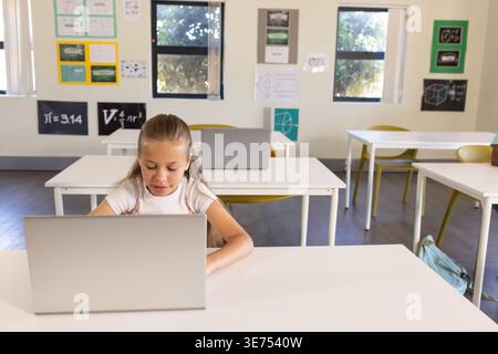 Child female student working on silver laptop at front desk in classroom with backpack. Girl, learner, education, school, daylight, posters, geometry Stock Photo