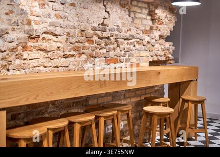 A long wooden bar counter with matching stools stands against an exposed rustic brick wall in a modern interior space. The light oak furniture Stock Photo