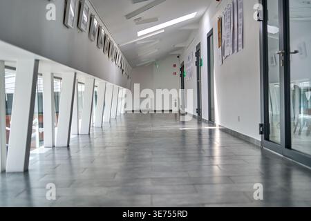 A long, modern, empty corridor in a contemporary public building featuring gray tiled flooring and white walls. The left side displays angled white Stock Photo