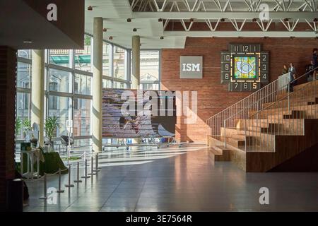 Interior view of a modern building lobby with high ceilings and exposed white trusses. A large red brick wall features an 'ISM' sign and a decorative Stock Photo