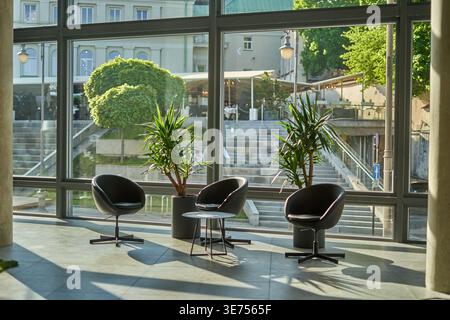 A modern interior lounge area featuring three black swivel armchairs and a small round table positioned on large stone tiles near floor-to-ceiling Stock Photo