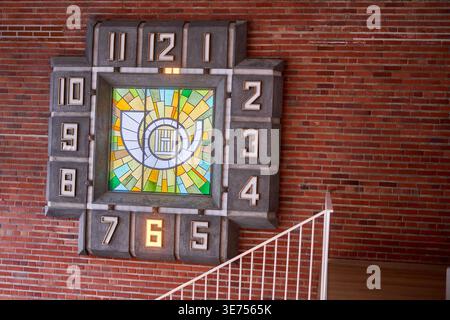 A large, vintage Art Deco style wall clock mounted on a red brick interior wall. The clock features a central square stained glass panel with Stock Photo