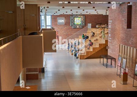 Interior view of a modern public building lobby featuring a large wooden amphitheater-style staircase with white railings where numerous people are Stock Photo