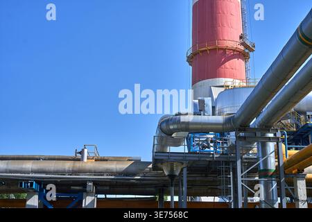 Industrial facility with extensive metal piping networks and a prominent red and white chimney stack against clear blue sky. Large silver pipelines Stock Photo